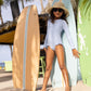 A young girl in a sunhat and sunglasses stands on sand between two surfboards, wearing the Snapper Rock Girls' Seaside Gingham Leg Frill Surf Suit with UPF50+ sun protection. Palm trees and a beach hut complete the scene.
