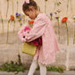 A young girl in a pink dress and white tights wears the Louise Misha Gilla Hair Clip as she stands with a basket of flowers against a floral-decorated wall.