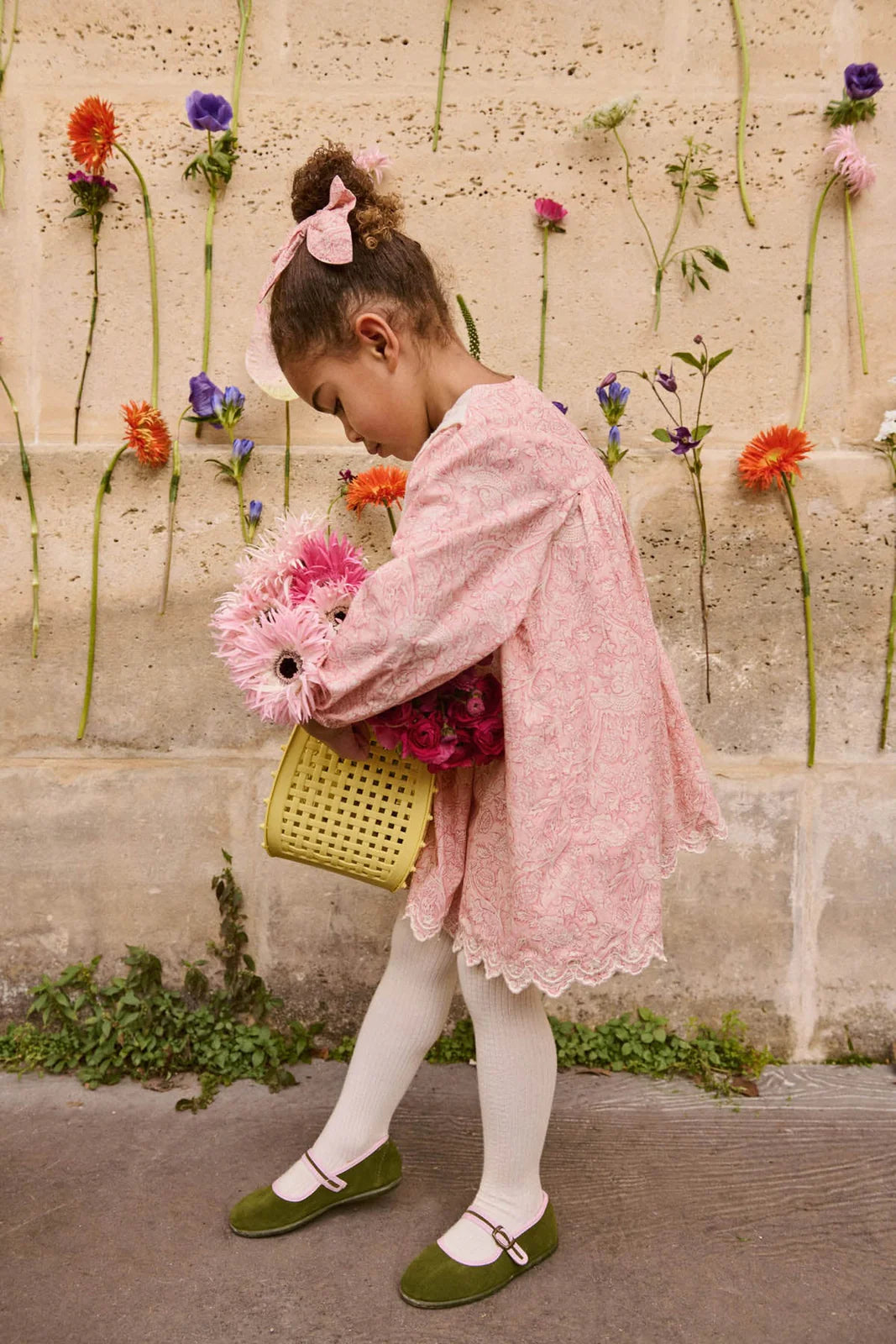A young girl in a pink dress and white tights wears the Louise Misha Gilla Hair Clip as she stands with a basket of flowers against a floral-decorated wall.