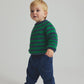 A young child stands on a white background, dressed in a green and navy striped sweater, Cozmo Baby Galo Denim Pants featuring blue herringbone twill fabric and an elastic waistband, paired with gray socks.