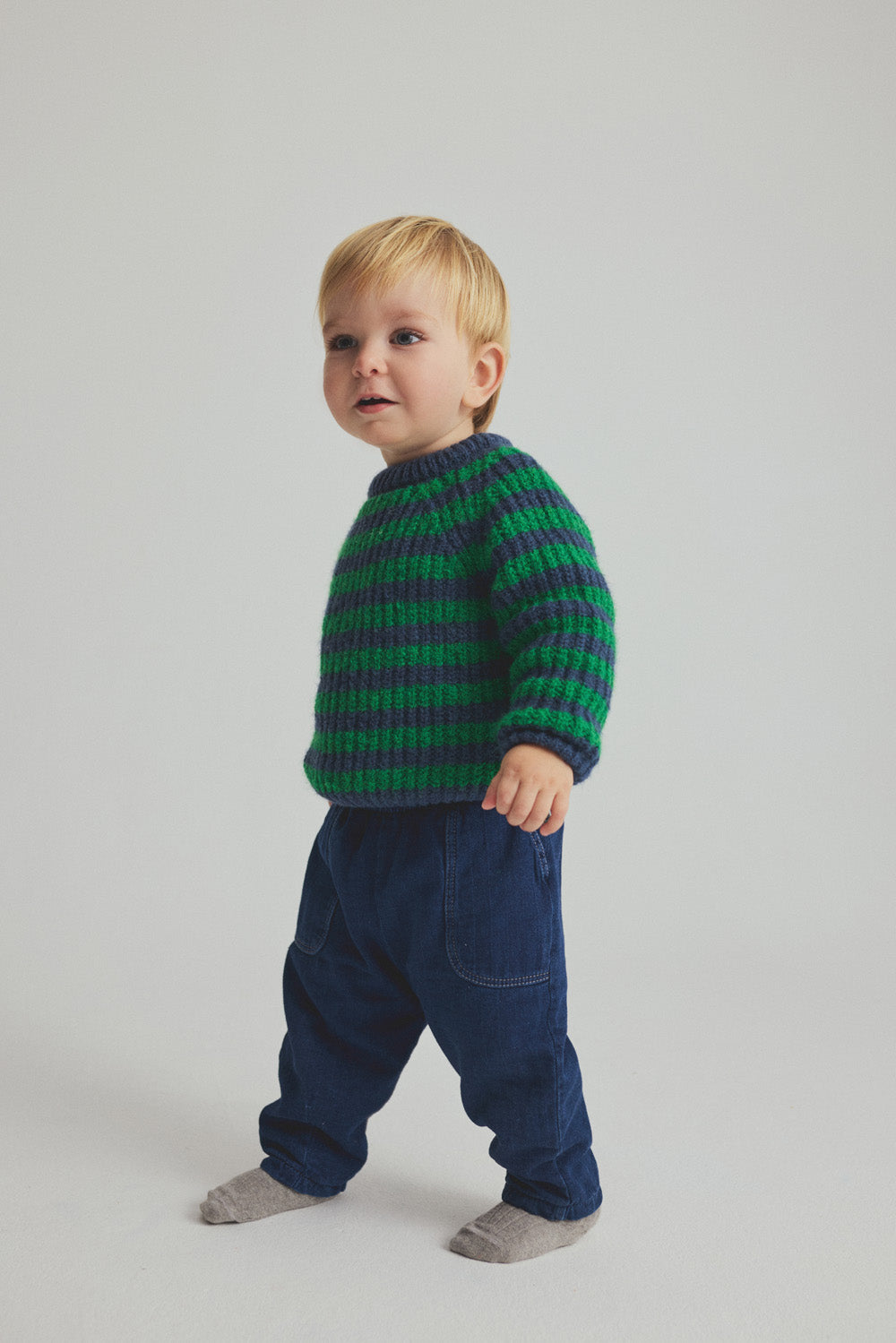 A young child stands on a white background, dressed in a green and navy striped sweater, Cozmo Baby Galo Denim Pants featuring blue herringbone twill fabric and an elastic waistband, paired with gray socks.