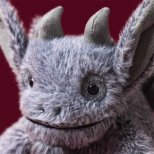 A close-up of the Jellycat Growlston Gargoyle plush toy—featuring soft gray faux fur, large round eyes, two small horns, and big pointed ears—set against a solid burgundy background.