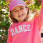 A young child smiles outdoors amid greenery and flowers, wearing the Bits & Bows Ballet Slippers Bow Hat and a pink “DANCE” sweatshirt.