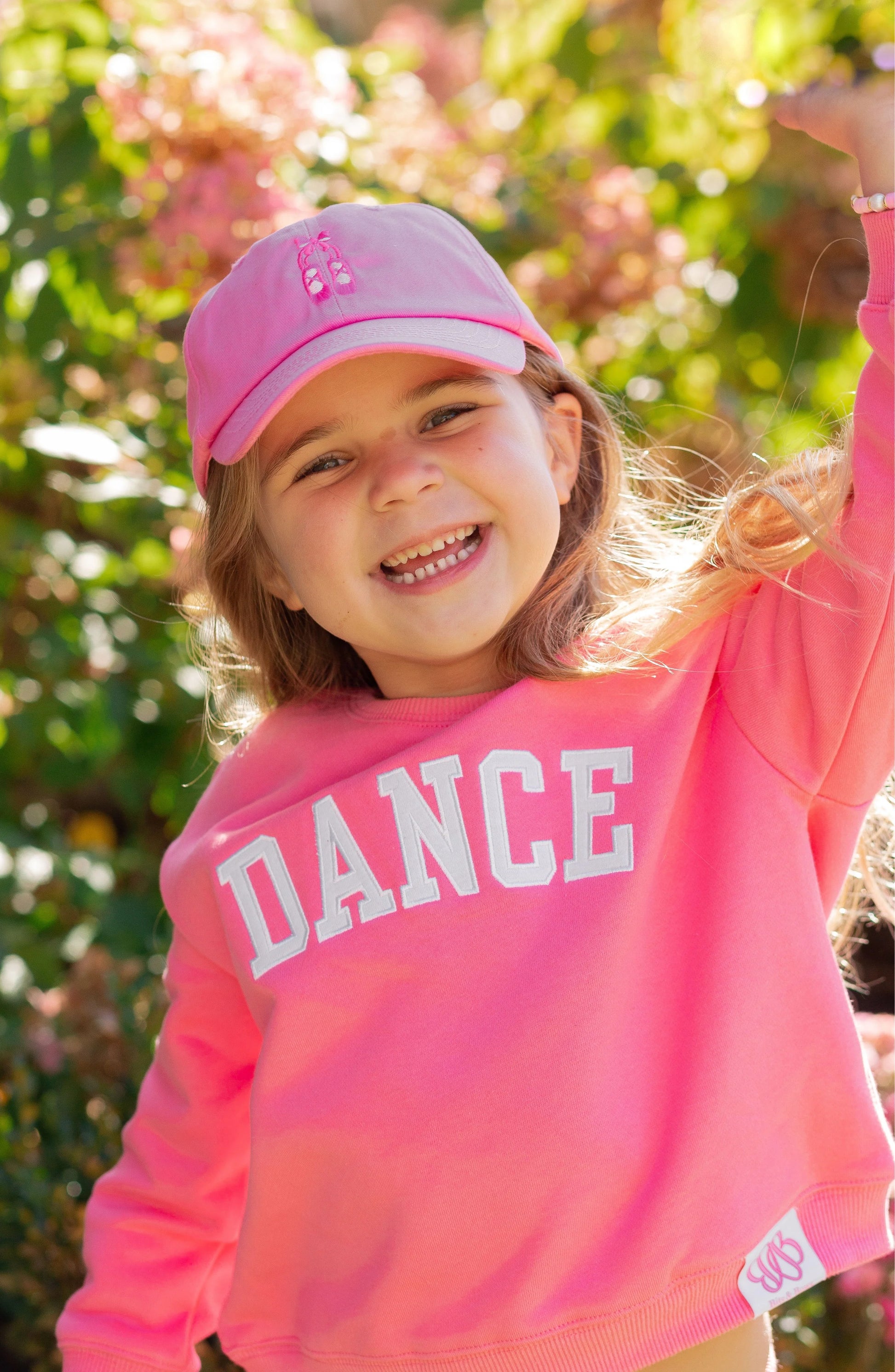A young child smiles outdoors amid greenery and flowers, wearing the Bits & Bows Ballet Slippers Bow Hat and a pink “DANCE” sweatshirt.