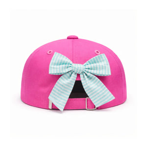 The Dolphin Daydream Bow Hat by Bits & Bows is a bright pink kids’ baseball cap, featuring a blue and white seersucker bow attached at the back, displayed on a white background.