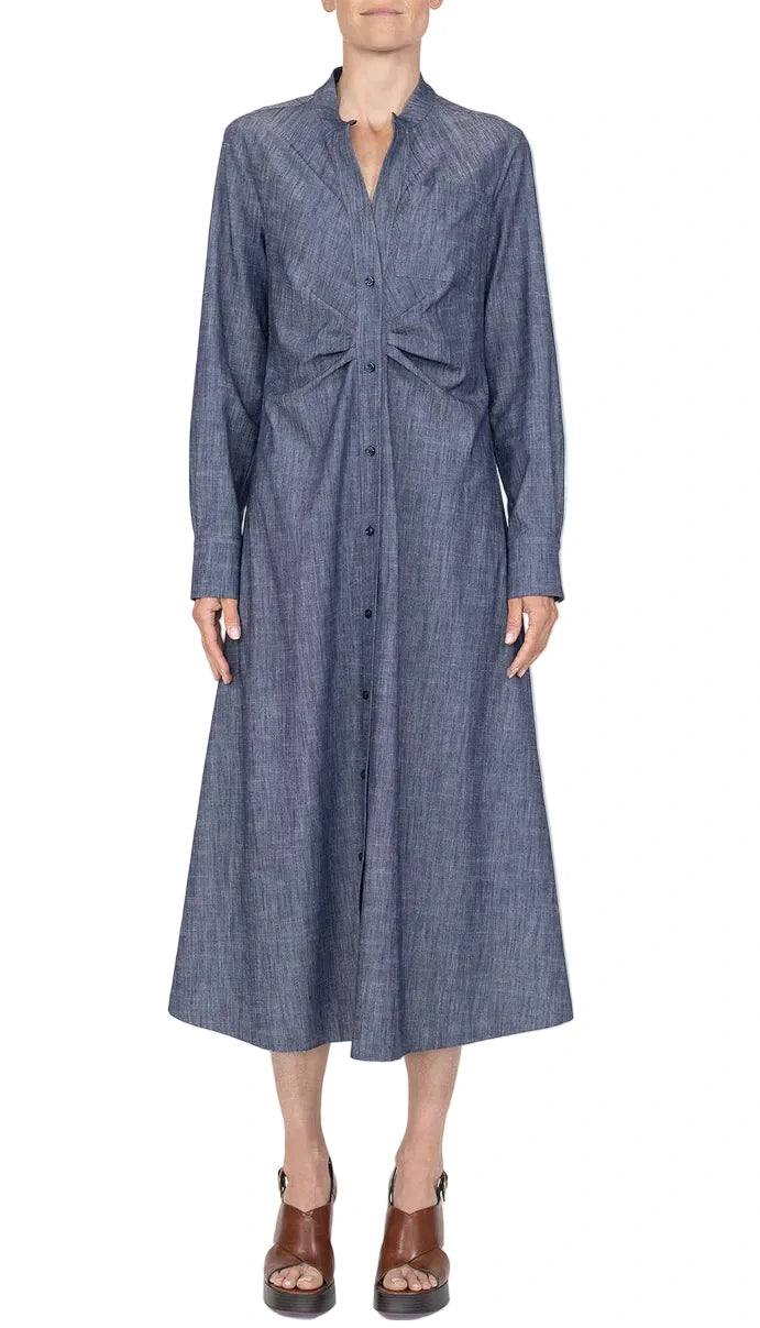 A woman wears the Genny Dress by Callas Milano—a blue chambray, long-sleeve, button-front piece with a fitted waist and classic fit-and-flare silhouette—styled with brown open-toe platform sandals.