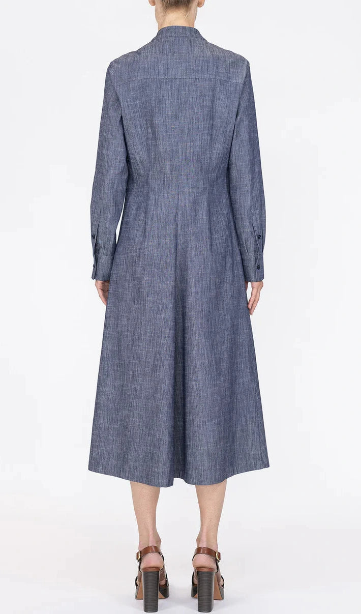 A person is shown from the back against a plain white background wearing the Callas Milano Genny Dress, a blue chambray long sleeve button-front style, paired with brown heeled sandals.