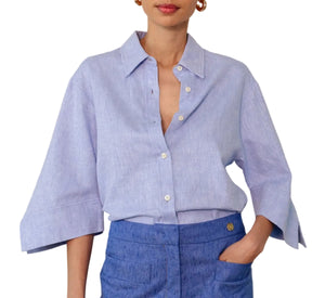 A woman wears the Georgie Top by Callas Milano, a classic light blue Italian twill shirt with a point collar and wide sleeves, tucked into high-waisted textured blue pants.