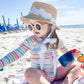 A young child in a striped swimsuit and Bits & Bows Youth Harbor Hat sits on a beach towel with sunglasses and toy cup, enjoying the sandy beach—offering fun style and sun protection. Blue chairs can be seen in the background.
