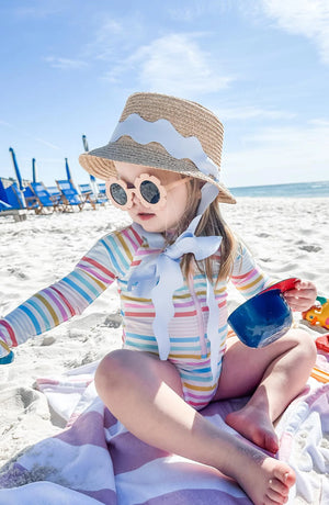 A young child in a striped swimsuit and Bits & Bows Youth Harbor Hat sits on a beach towel with sunglasses and toy cup, enjoying the sandy beach—offering fun style and sun protection. Blue chairs can be seen in the background.