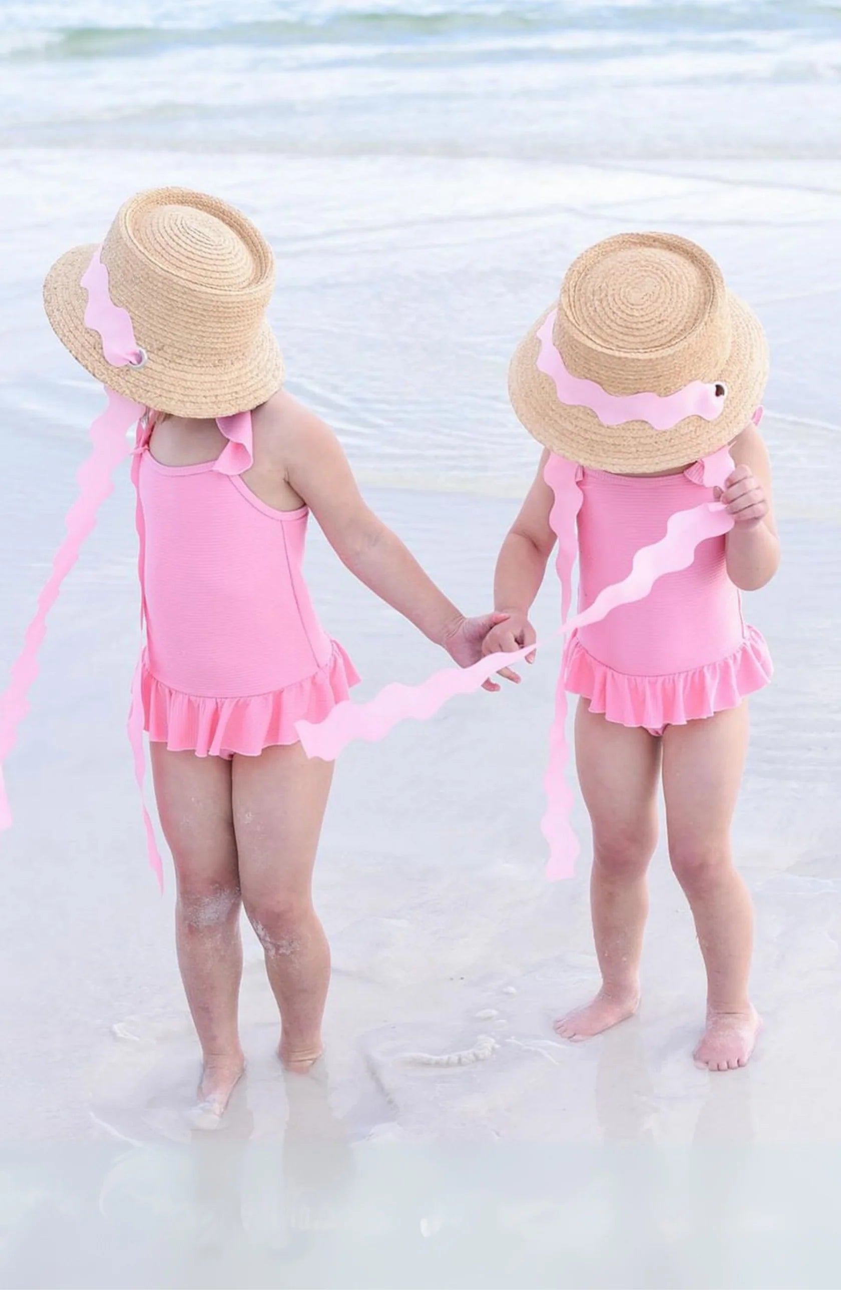Two young children in matching Youth Harbor Hats by Bits & Bows hold hands in shallow beach water, enjoying both style and sun protection.