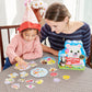 An adult and a child wearing a party hat enjoy quality family time as they play the Eeboo Cupcake Shaped Spinner Game, matching colorful pieces on a checkered tablecloth and spinning the cupcake-shaped spinner for extra fun.