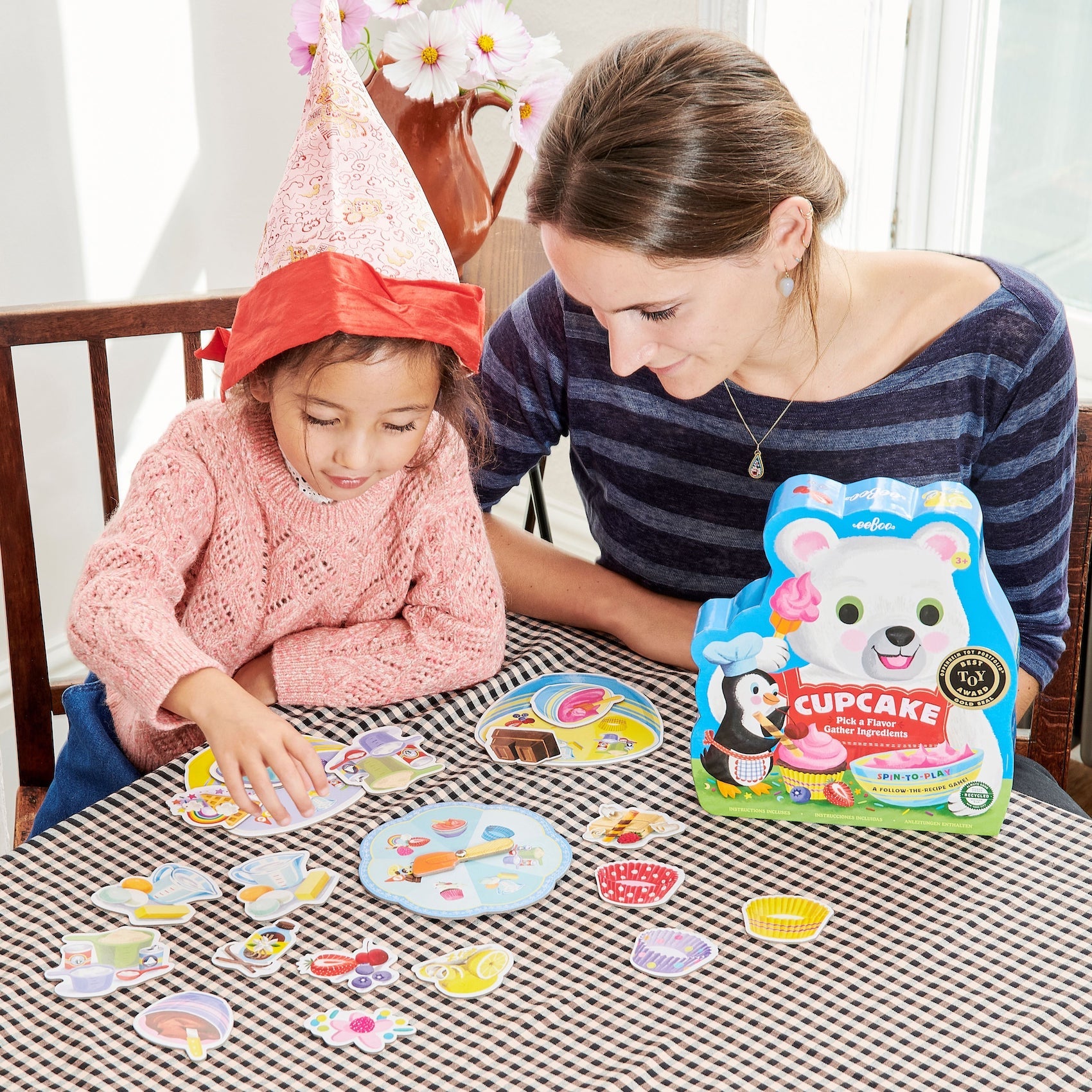 An adult and a child wearing a party hat enjoy quality family time as they play the Eeboo Cupcake Shaped Spinner Game, matching colorful pieces on a checkered tablecloth and spinning the cupcake-shaped spinner for extra fun.
