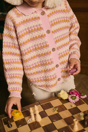 A child wearing the Louise Misha Girls' Khellani Cape stands by a chessboard, holding flowers and placing them among the chess pieces.