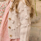 A child stands sideways against a stone wall, wearing Louise Misha's Baby Girls' and Girls' Sandy Cardigan—a cream knit piece with a buttoned front and textured hand-crocheted flowers—over a light pink patterned dress.