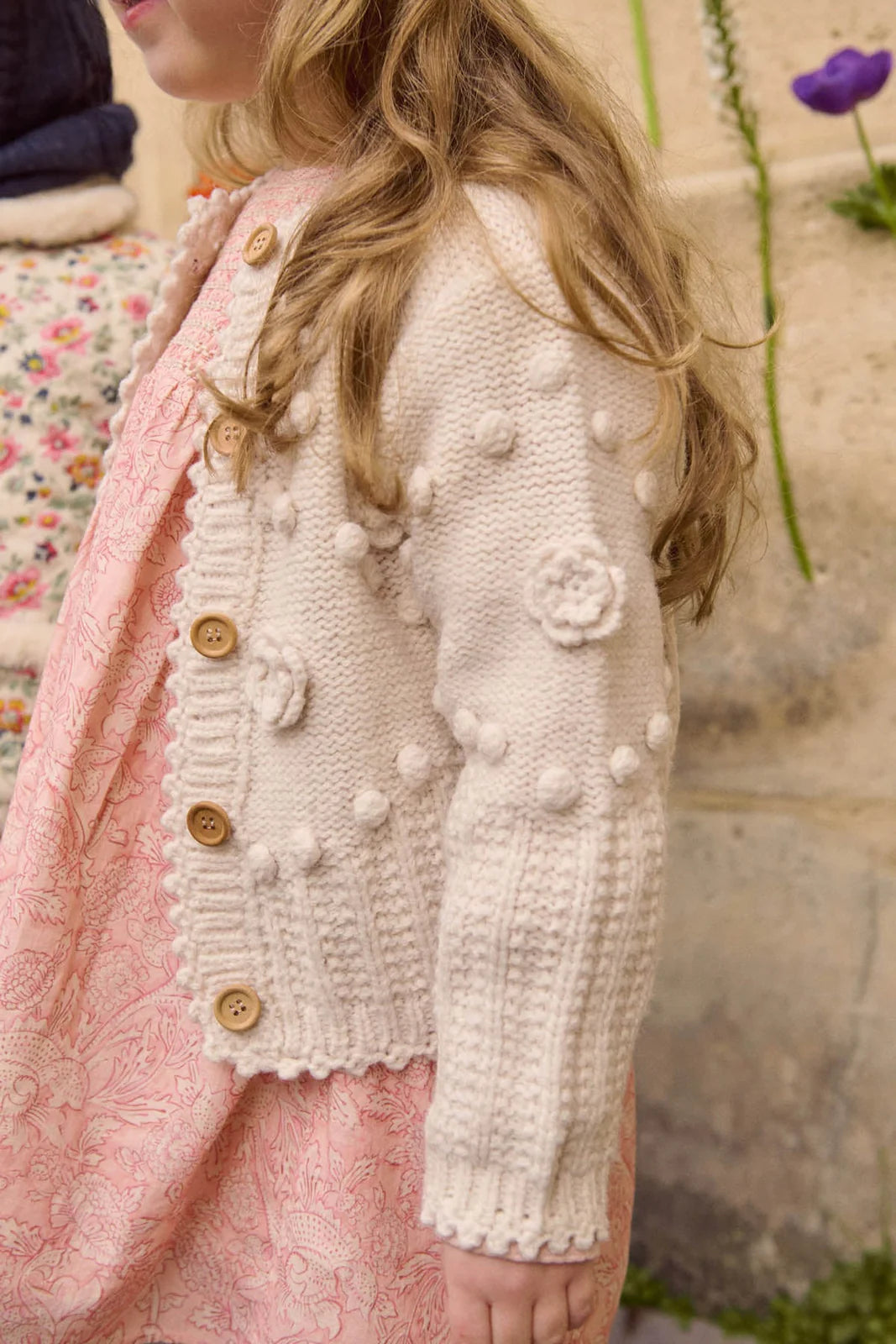 A child stands sideways against a stone wall, wearing Louise Misha's Baby Girls' and Girls' Sandy Cardigan—a cream knit piece with a buttoned front and textured hand-crocheted flowers—over a light pink patterned dress.