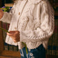 A young child wears the Louise Misha Girls' Khellana Cape Sweater, a hand-crocheted white cardigan with a Peter Pan collar, along with blue jeans featuring white embroidery, while holding a green flower stem.