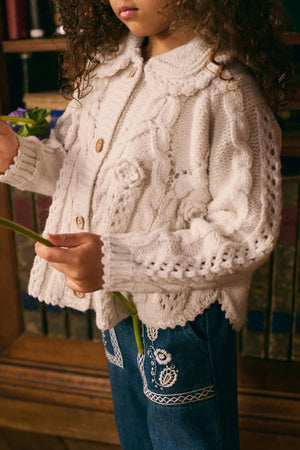 A young child wears the Louise Misha Girls' Khellana Cape Sweater, a hand-crocheted white cardigan with a Peter Pan collar, along with blue jeans featuring white embroidery, while holding a green flower stem.