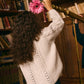 A child with curly hair wearing the Louise Misha Girls' Khellana Cape Sweater holds a pink flower and looks through a brass telescope in a book-filled library.