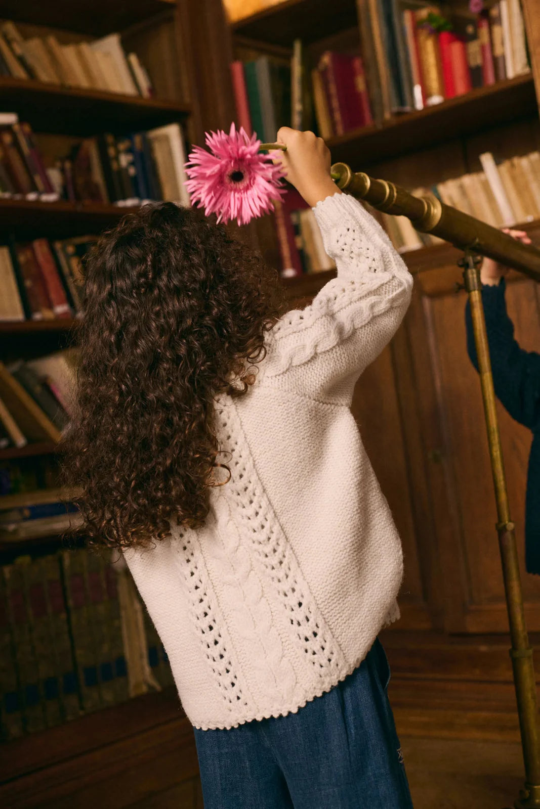 A child with curly hair wearing the Louise Misha Girls' Khellana Cape Sweater holds a pink flower and looks through a brass telescope in a book-filled library.