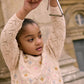 A young girl wears the Louise Misha Girls' Jadeli Jumper, a cream-colored knit sweater with a floral pattern, while holding a metal triangle instrument above her head outdoors.