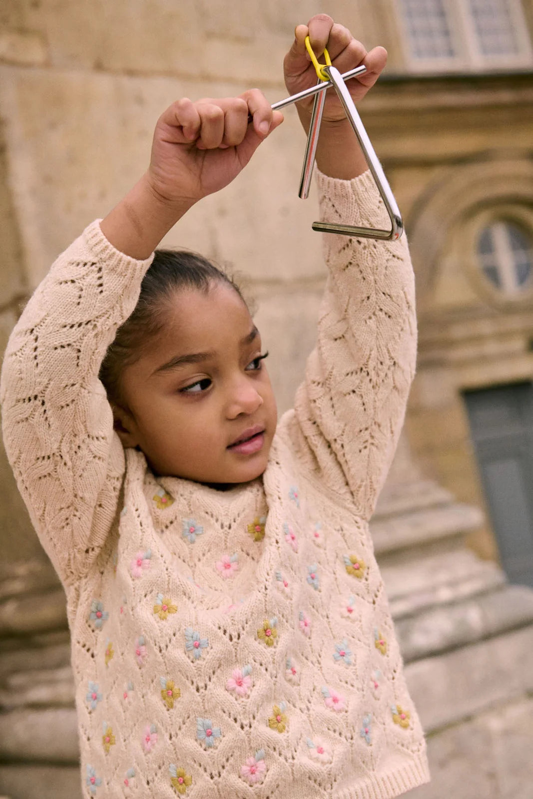 A young girl wears the Louise Misha Girls' Jadeli Jumper, a cream-colored knit sweater with a floral pattern, while holding a metal triangle instrument above her head outdoors.