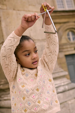 A young girl wears the Louise Misha Girls' Jadeli Jumper, a cream-colored knit sweater with a floral pattern, while holding a metal triangle instrument above her head outdoors.