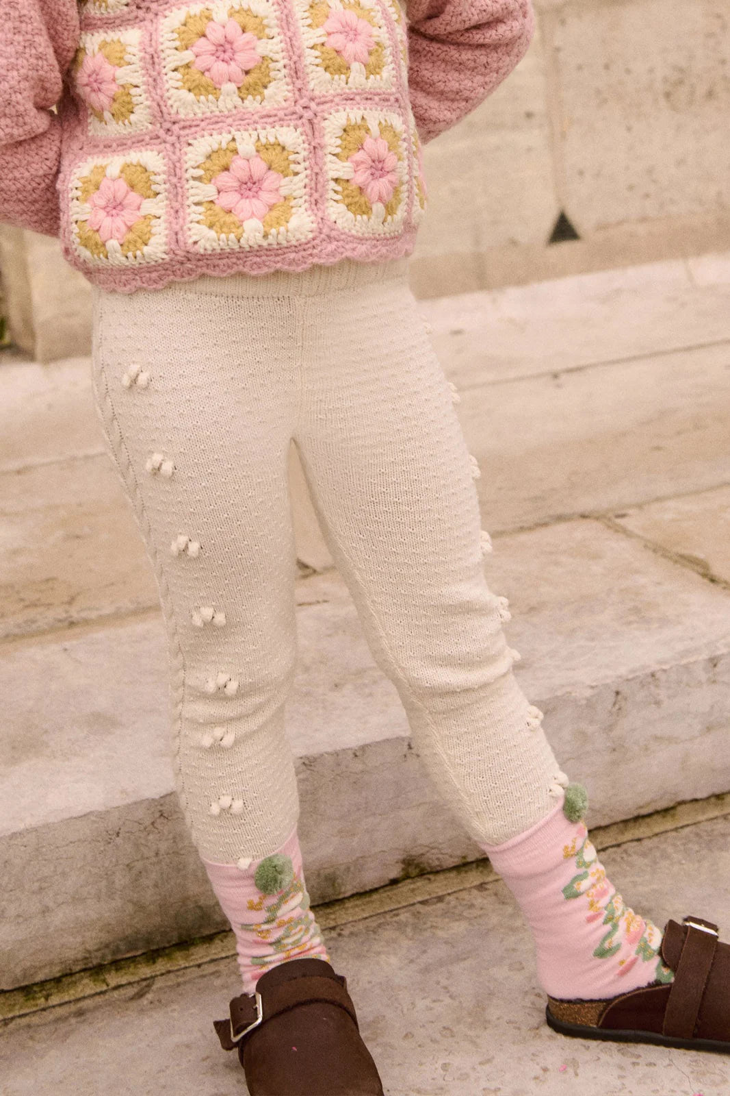 A child wearing Louise Misha Baby Girls' and Girls' Molda Leggings with an elasticized waist stands on stone steps, paired with pink and green socks, brown sandals, and a pink crochet sweater.