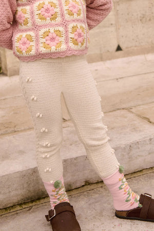 A child wearing Louise Misha Baby Girls' and Girls' Molda Leggings with an elasticized waist stands on stone steps, paired with pink and green socks, brown sandals, and a pink crochet sweater.