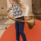 A young girl stands on an orange platform, holding a large brass horn, wearing a headband, blue pants, brown shoes, and the Louise Misha Girls' Rynia Crochet Top.