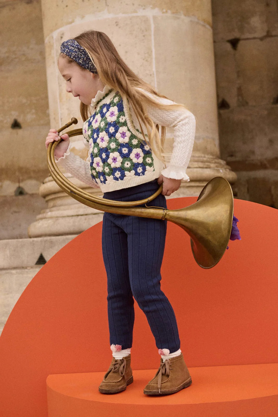 A young girl stands on an orange platform, holding a large brass horn, wearing a headband, blue pants, brown shoes, and the Louise Misha Girls' Rynia Crochet Top.