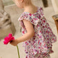 A young child wearing the Louise Misha Girls’ Inessa Blouse, featuring a floral cotton design with a ruffled lace square neckline, holds a bright pink flower while walking indoors.