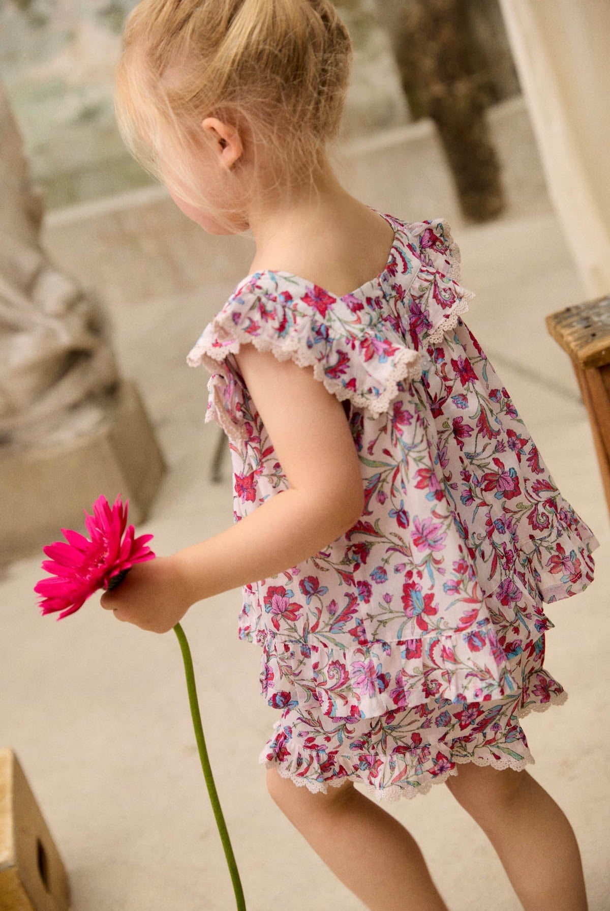 A young child wearing the Louise Misha Girls’ Inessa Blouse, featuring a floral cotton design with a ruffled lace square neckline, holds a bright pink flower while walking indoors.