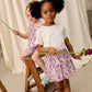 Two young girls in floral Louise Misha outfits—one wearing a delicate cotton veil and the other in the Girls’ Roumia Skirt featuring a smocked waistband—hold long-stem flowers as they stand and sit on a wooden ladder against a draped fabric backdrop.