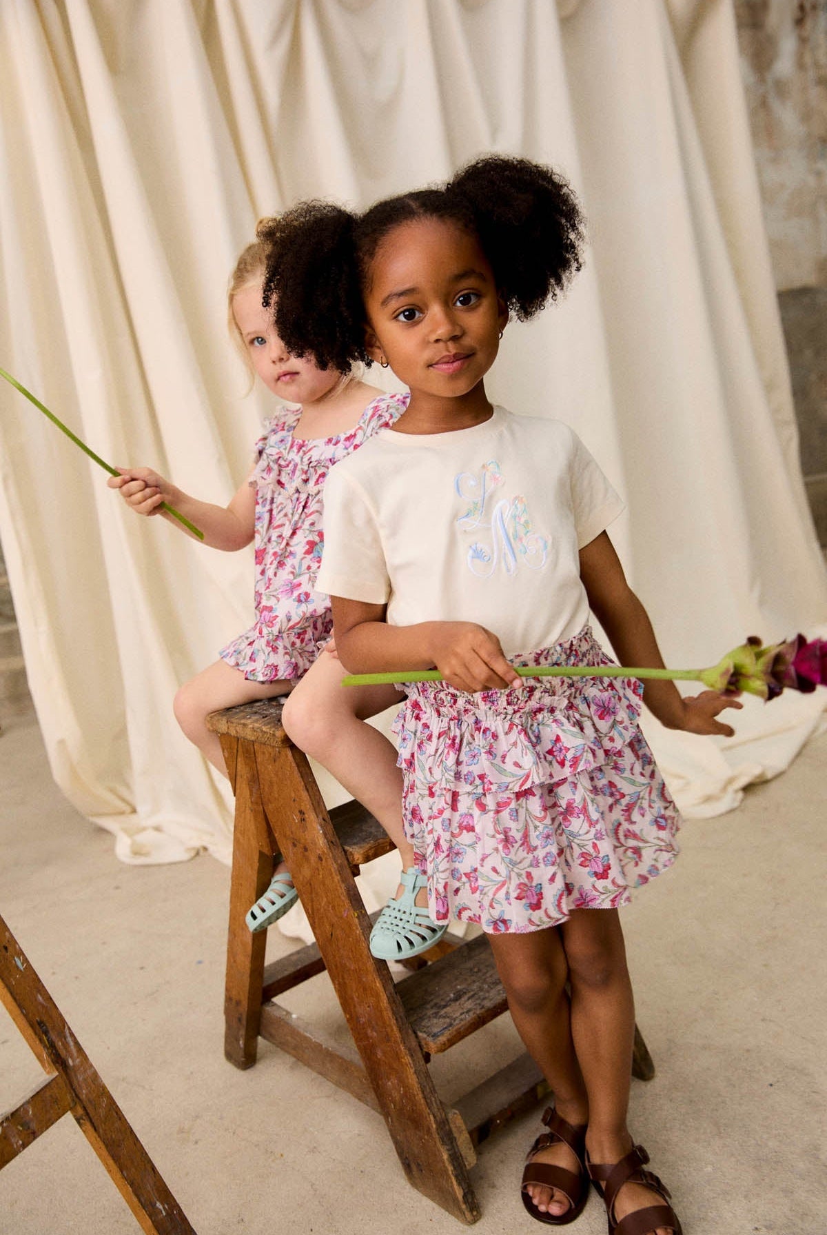 Two young girls in floral Louise Misha outfits—one wearing a delicate cotton veil and the other in the Girls’ Roumia Skirt featuring a smocked waistband—hold long-stem flowers as they stand and sit on a wooden ladder against a draped fabric backdrop.