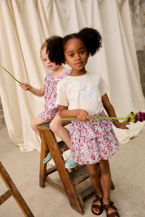 Two young girls in floral Louise Misha outfits—one wearing a delicate cotton veil and the other in the Girls’ Roumia Skirt featuring a smocked waistband—hold long-stem flowers as they stand and sit on a wooden ladder against a draped fabric backdrop.
