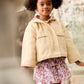 A young girl with puffy hair wears a light jacket over the Louise Misha Girls’ Roumia Skirt, which features a smocked waistband and a floral print, standing indoors near sculptures and a weathered wall.