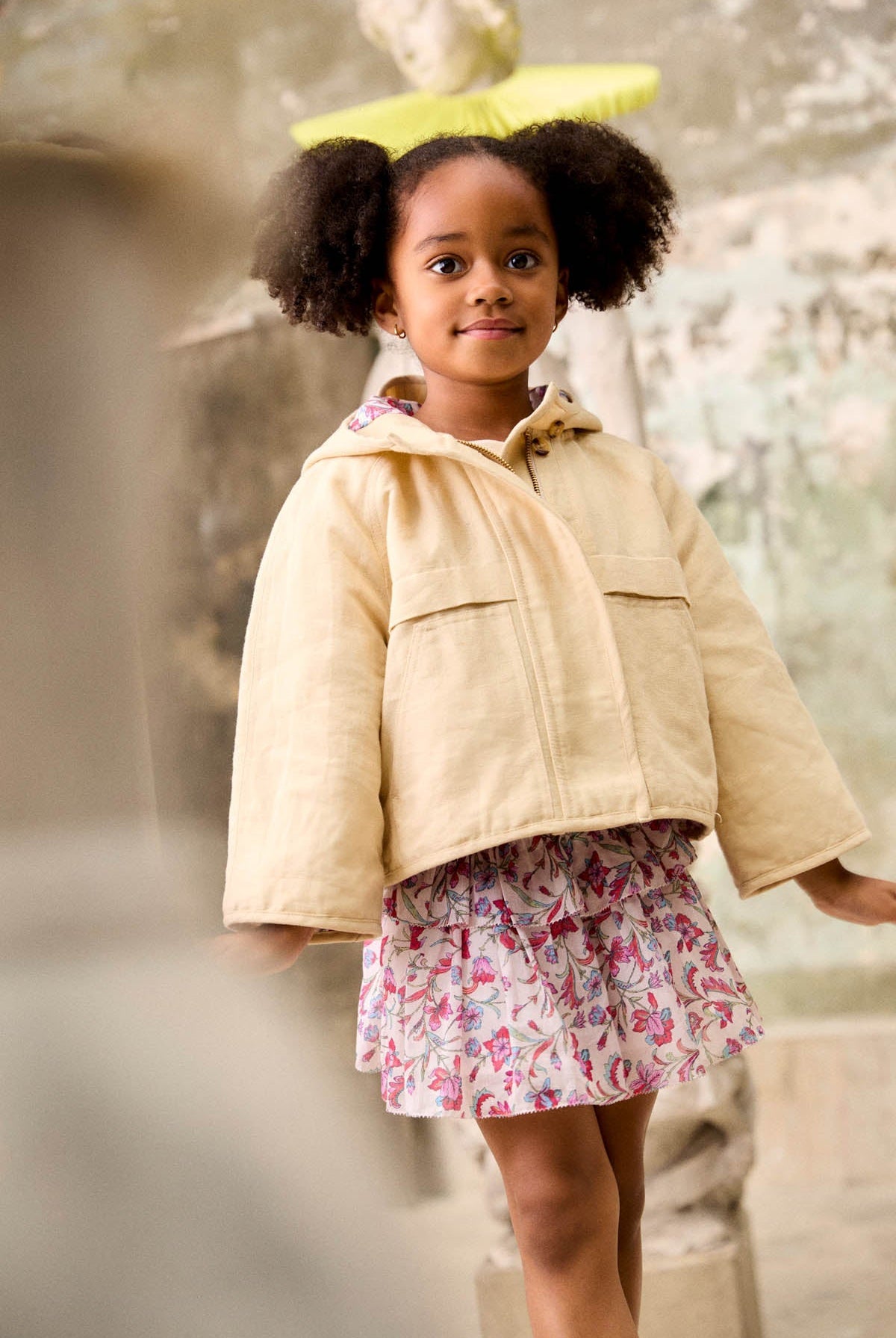 A young girl with puffy hair wears a light jacket over the Louise Misha Girls’ Roumia Skirt, which features a smocked waistband and a floral print, standing indoors near sculptures and a weathered wall.