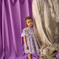 A young girl in the Louise Misha Girls’ Siloe Dress, featuring a floral cotton pattern and ruffled sleeves, stands beside a marble statue with a purple draped curtain and weathered wall in the background.