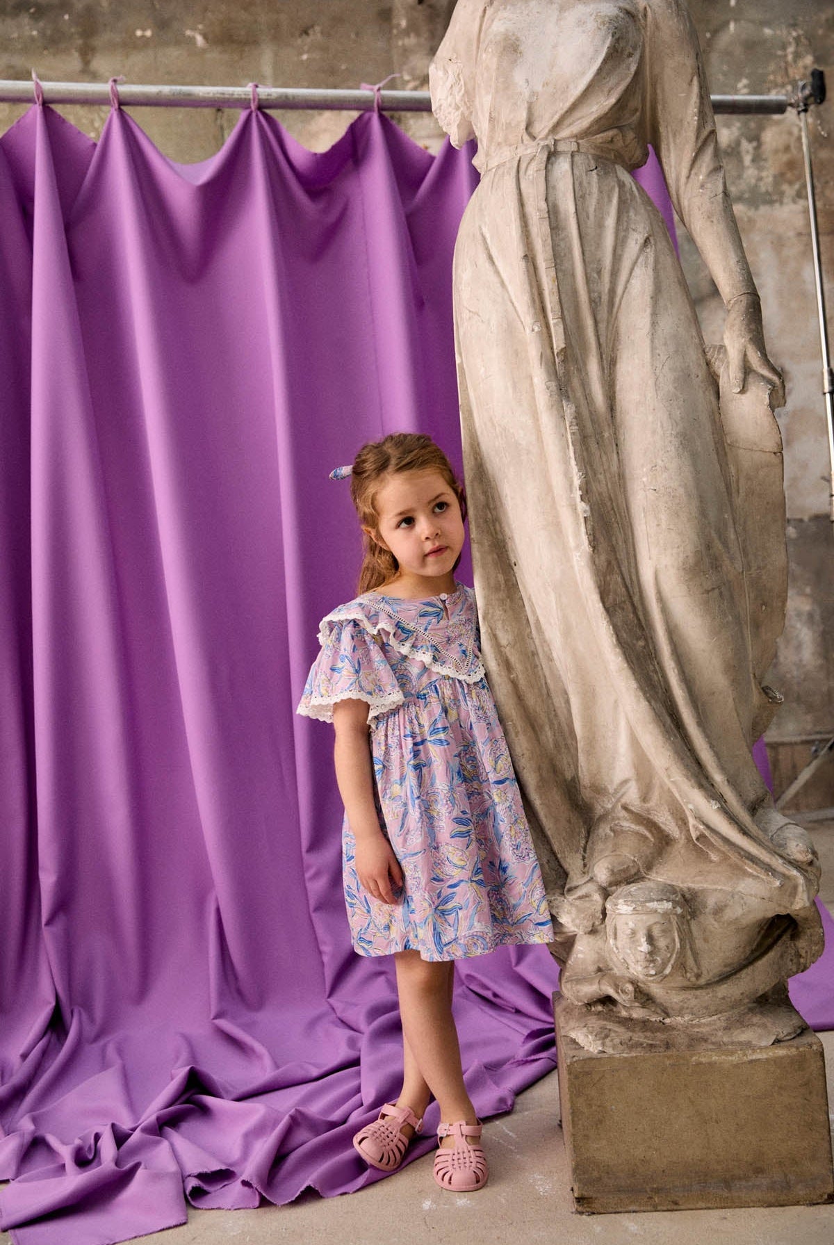 A young girl in the Louise Misha Girls’ Siloe Dress, featuring a floral cotton pattern and ruffled sleeves, stands beside a marble statue with a purple draped curtain and weathered wall in the background.