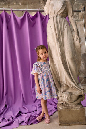 A young girl in the Louise Misha Girls’ Siloe Dress, featuring a floral cotton pattern and ruffled sleeves, stands beside a marble statue with a purple draped curtain and weathered wall in the background.