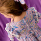 A young girl wearing the Louise Misha Girls’ Siloe Dress, featuring floral cotton fabric with ruffled sleeves and lace trim, along with a matching hair scrunchie, stands in front of a purple backdrop.