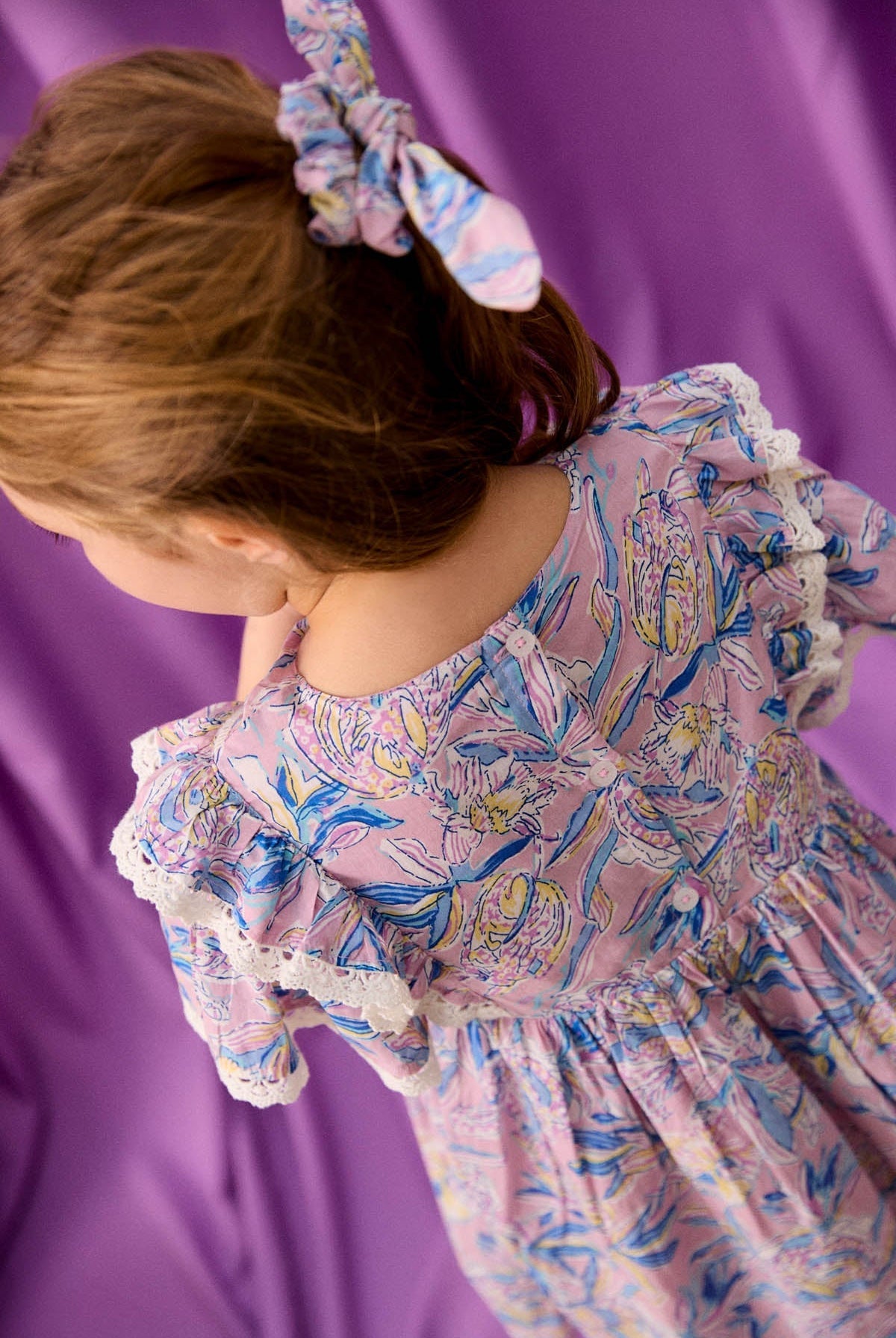 A young girl wearing the Louise Misha Girls’ Siloe Dress, featuring floral cotton fabric with ruffled sleeves and lace trim, along with a matching hair scrunchie, stands in front of a purple backdrop.