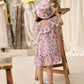 A young child wearing the Louise Misha Girls’ Natiyana Dress—a floral cotton dress with ruffled sleeves—and a hat stands by a wooden stand, gazing at a stone bust in an art studio filled with sculptures and a ladder in the background.