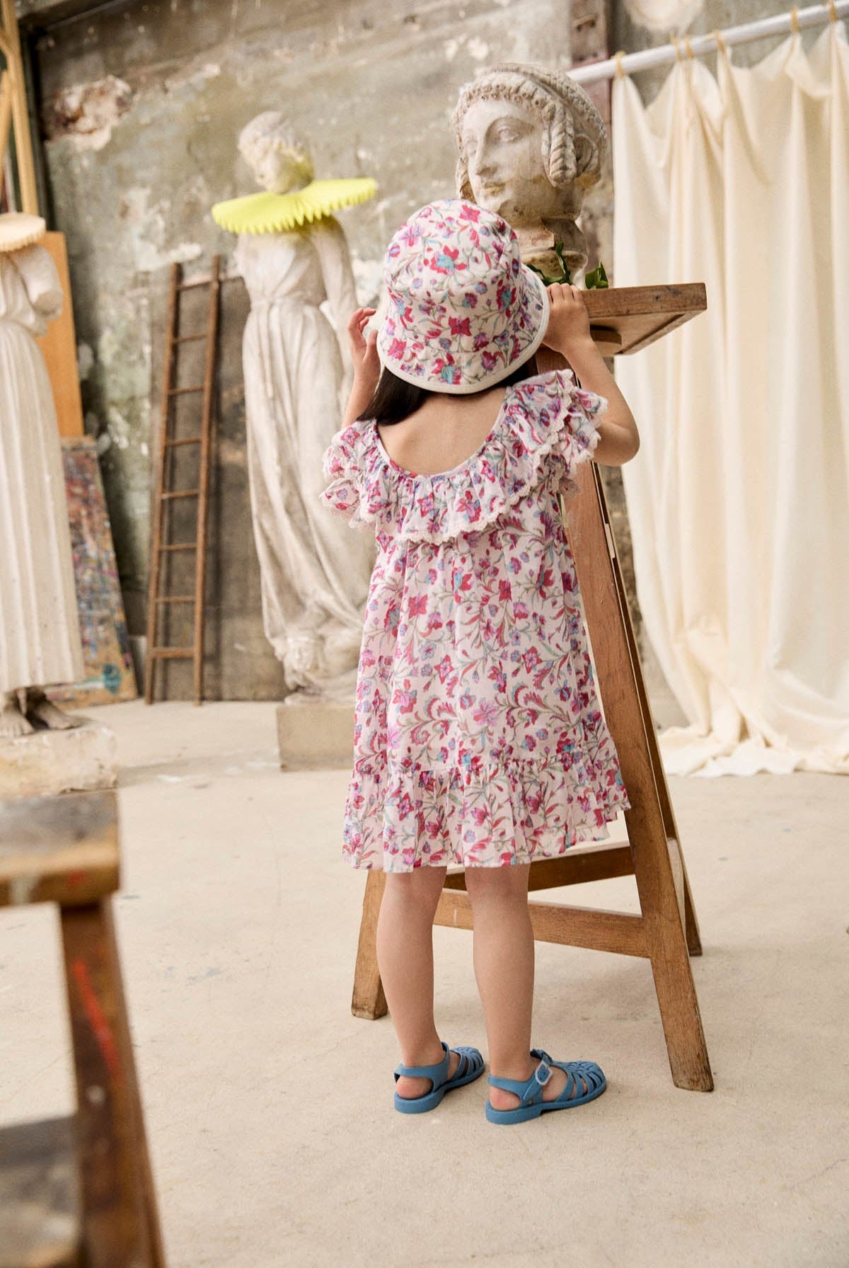 A young child wearing the Louise Misha Girls’ Natiyana Dress—a floral cotton dress with ruffled sleeves—and a hat stands by a wooden stand, gazing at a stone bust in an art studio filled with sculptures and a ladder in the background.