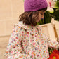A young child in the Louise Misha Girls' Cleo Blouse and a purple knit hat stands next to a crate of assorted flowers.