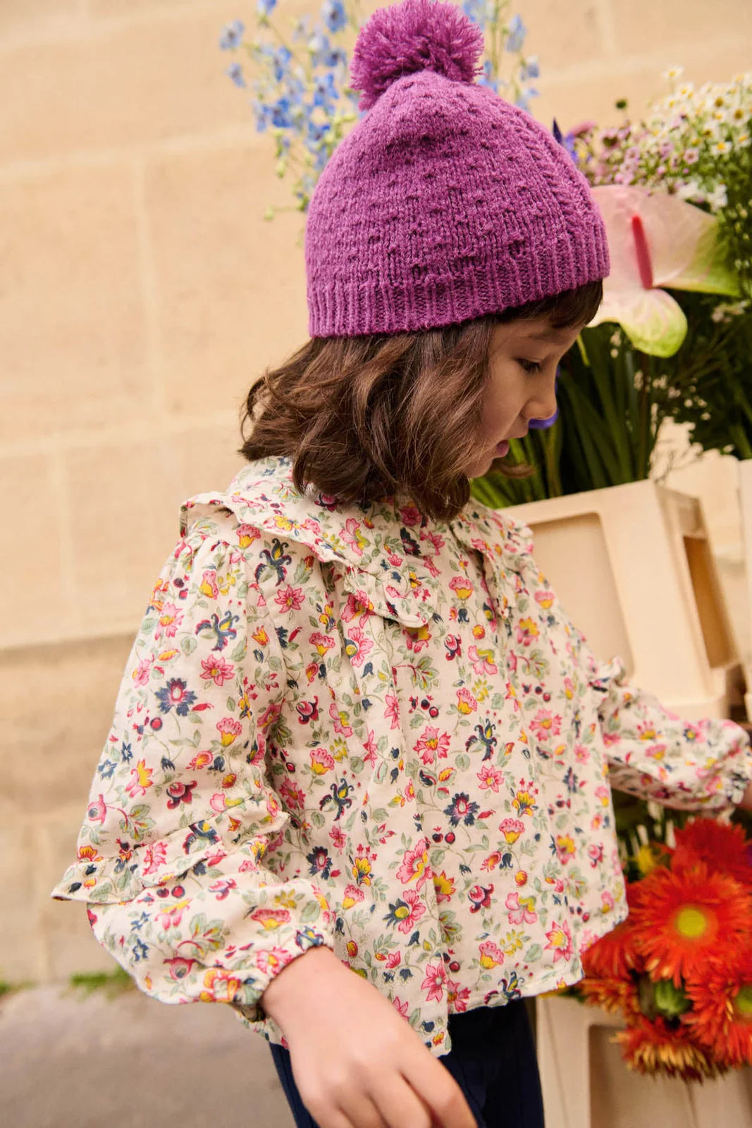 A young child in the Louise Misha Girls' Cleo Blouse and a purple knit hat stands next to a crate of assorted flowers.