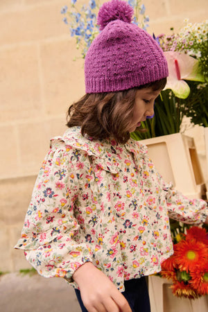 A young child in the Louise Misha Girls' Cleo Blouse and a purple knit hat stands next to a crate of assorted flowers.