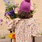 A child in a purple knit hat and the Louise Misha Girls' Cleo Blouse stands with her back to the camera, holding a flower amid vibrant blooms.