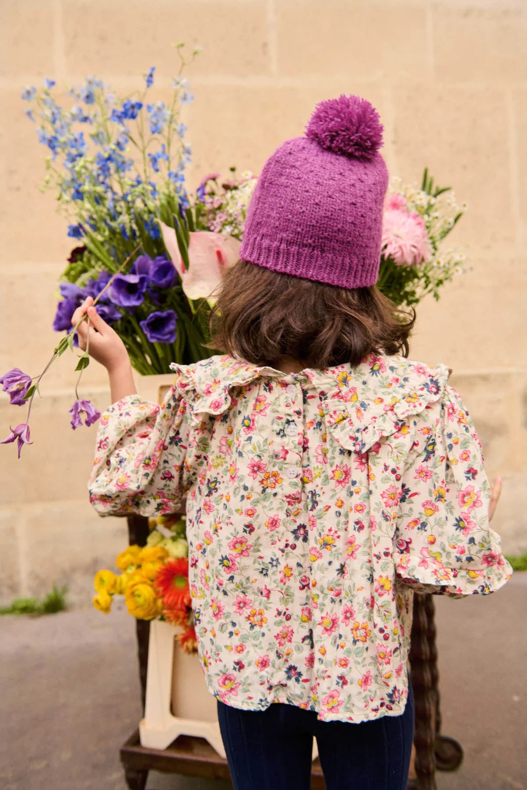 A child in a purple knit hat and the Louise Misha Girls' Cleo Blouse stands with her back to the camera, holding a flower amid vibrant blooms.